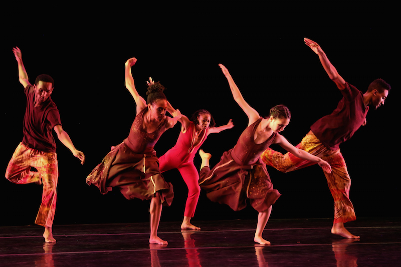 A group of five dancers wearing shades of red stand on one leg. Their arms are outstretched like wings and their other leg is bent behind them.